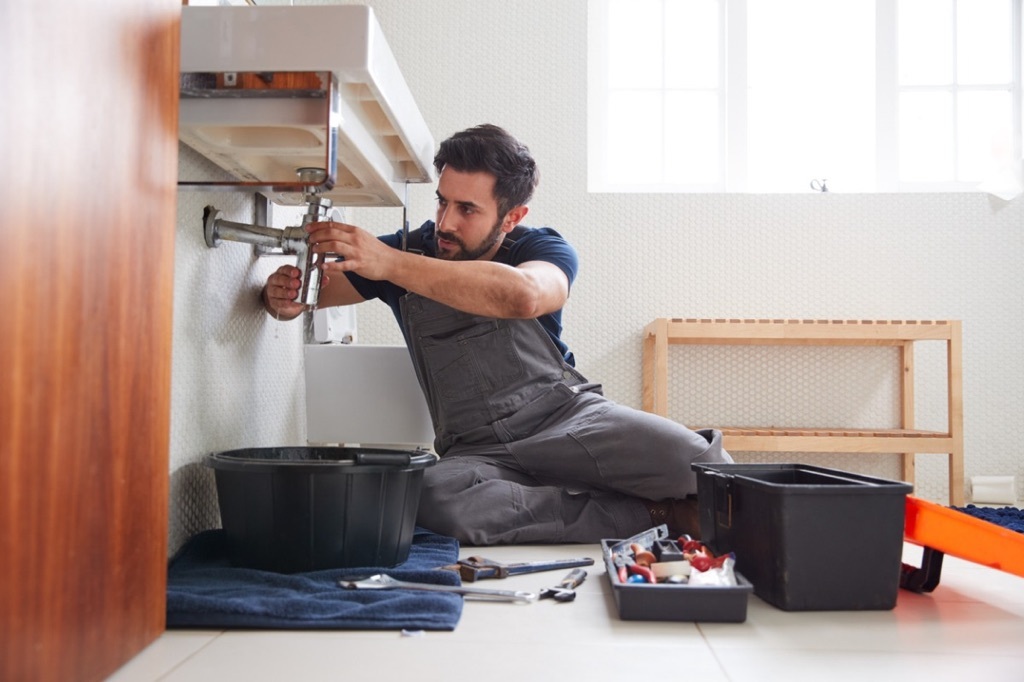 Person sitting on the floor under a sink fixing pipes, with tools around on the floor.